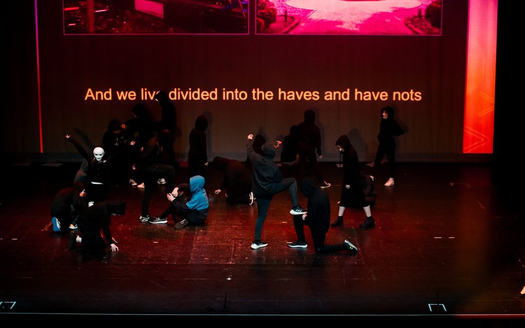 A group of young people performing onstage dressed in black with the words 'we live divided into the haves and have nots' behind them