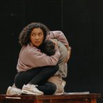 A woman with brown curly hair crouched down to hold a man kneeling. She is wearing a pink jumper and he is wearing a green jumper. They are both on top of a wooden table.