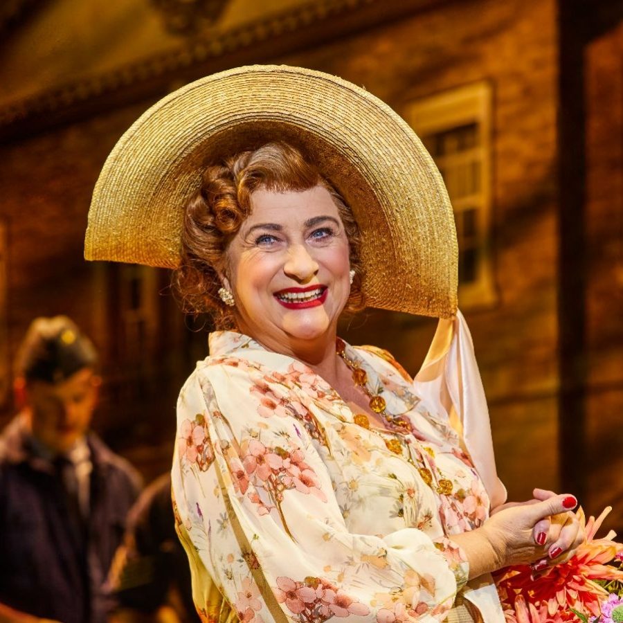 Caroline Quentin as Mrs Malaprop, wearing a wide-brimmed hat and a big smile, holding a bunch of flowers, with airmen and a country house in the background.