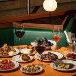 On a table, a selection of small plates of Italian dishes and glasses filled with wine are spread out.