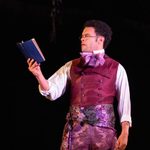 An actor on stage, dressed in a purple waistcoat and white shirt, gazes thoughtfully at a blue book he is holding up
