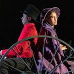 Actors in period costumes on a stage carriage, one in a red shirt and top hat driving, and the other in a purple dress and bonnet, with focused expressions.