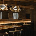 LASDUN Restaurant bar, with stools, and interior with table settings, large pendant lights and mirrors.