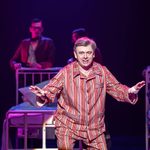 Michael Sheen wearing red striped pyjamas. He is mid-song. The ensemble cast move hospital beds behind him.