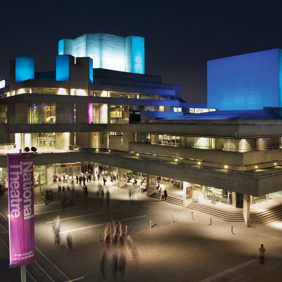 The exterior of the National Theatre at night, with the flytowers lit up in blue light.