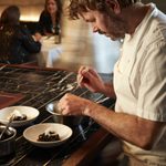 A chef stands at the bar plating up a squid ink and cuttlefish dish into a white bowl. The bar is black marble tile, the chef has a medium beard, floppy blonde hair, he wears chef whites.