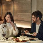 A woman with dark lond har sits on the left, she is laughing and is holding her hand up to her mouth mid laugh. She is sat next to a man with brown hair, a light beard and glasses who is looking at her. They are sharing a steak.