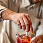 A bartenders hands are finishing a negroni in a tumble. The liquid is a light red, his hands are rubbing orange rind around the rim. he wears a service jacket in cream with a logo on the breast pocket.