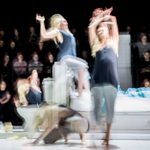 A blurred photograph of four figures in various poses, all with blond hair and dressed identically, on a stage set representing a bathroom and bedroom, and an audience is beyond them.