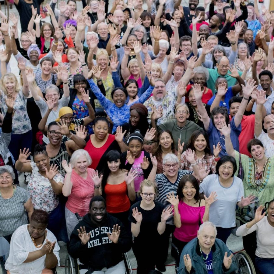 A large crowd of people stand waving at the camera. The crowd includes people from a diverse range of ages, accessibility needs and ethnicities.