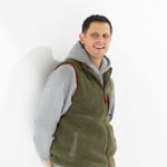 A smiling man wearing a gilet over a hoodie stands, leaning agaist a white wall.