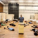 The cast of Nye performing in a rehearsal room. A man stands in the middle of the room and thirteen people lie on the floor around him.